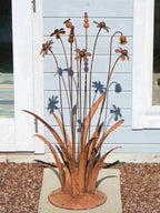A rusty steel meadow composition - poppies, chamomiles, heleniums and reeds rising through tall grasses on a hand-formed domed base, in front of a blue wooden cabin