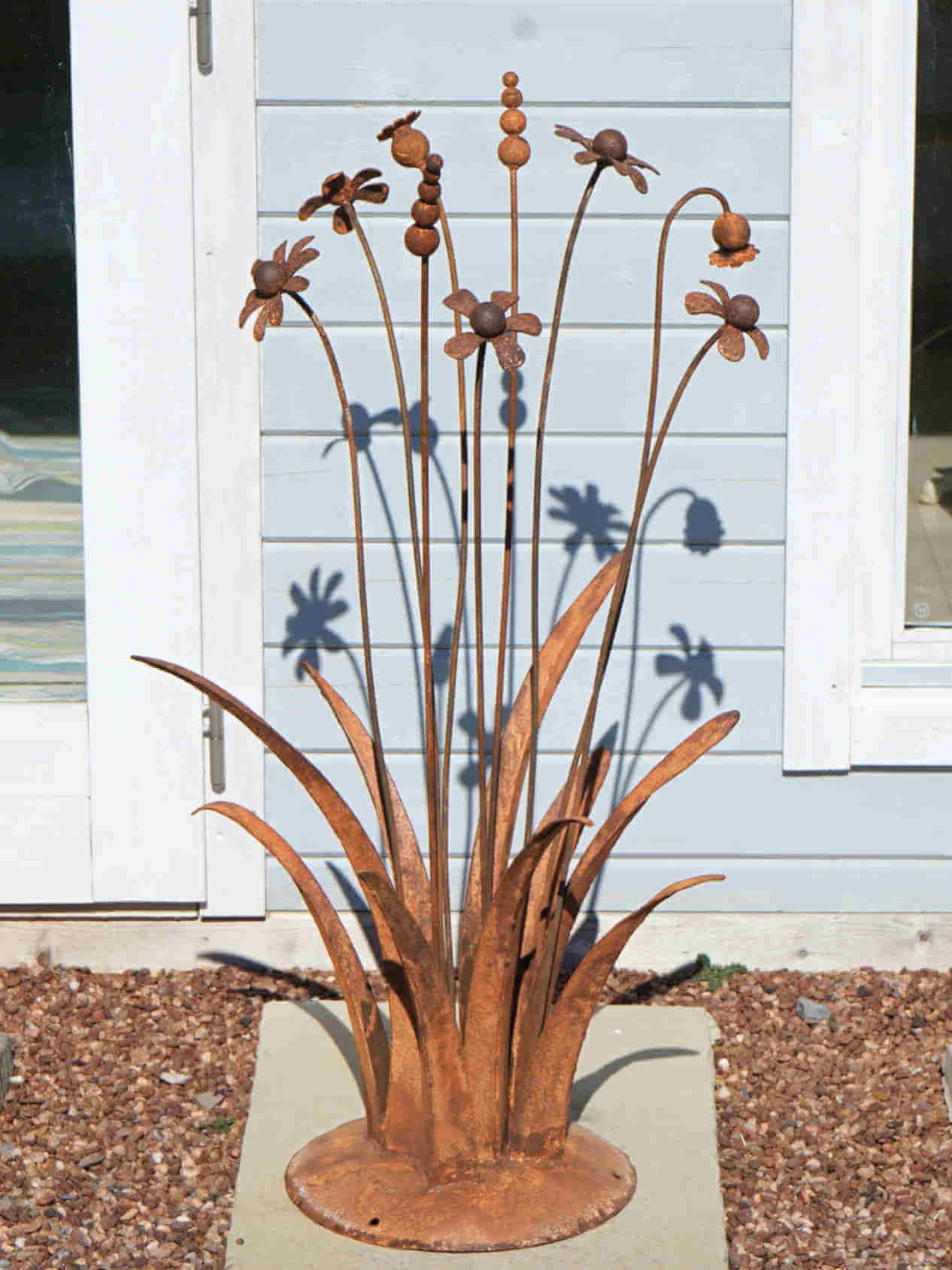 A rusty steel meadow composition - poppies, chamomiles, heleniums and reeds rising through tall grasses on a hand-formed domed base, in front of a blue wooden cabin