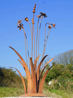 A rusty steel meadow composition - poppies, chamomiles, heleniums and reeds rising through tall grasses on a hand-formed domed base, with a blue sky and greenery in the background
