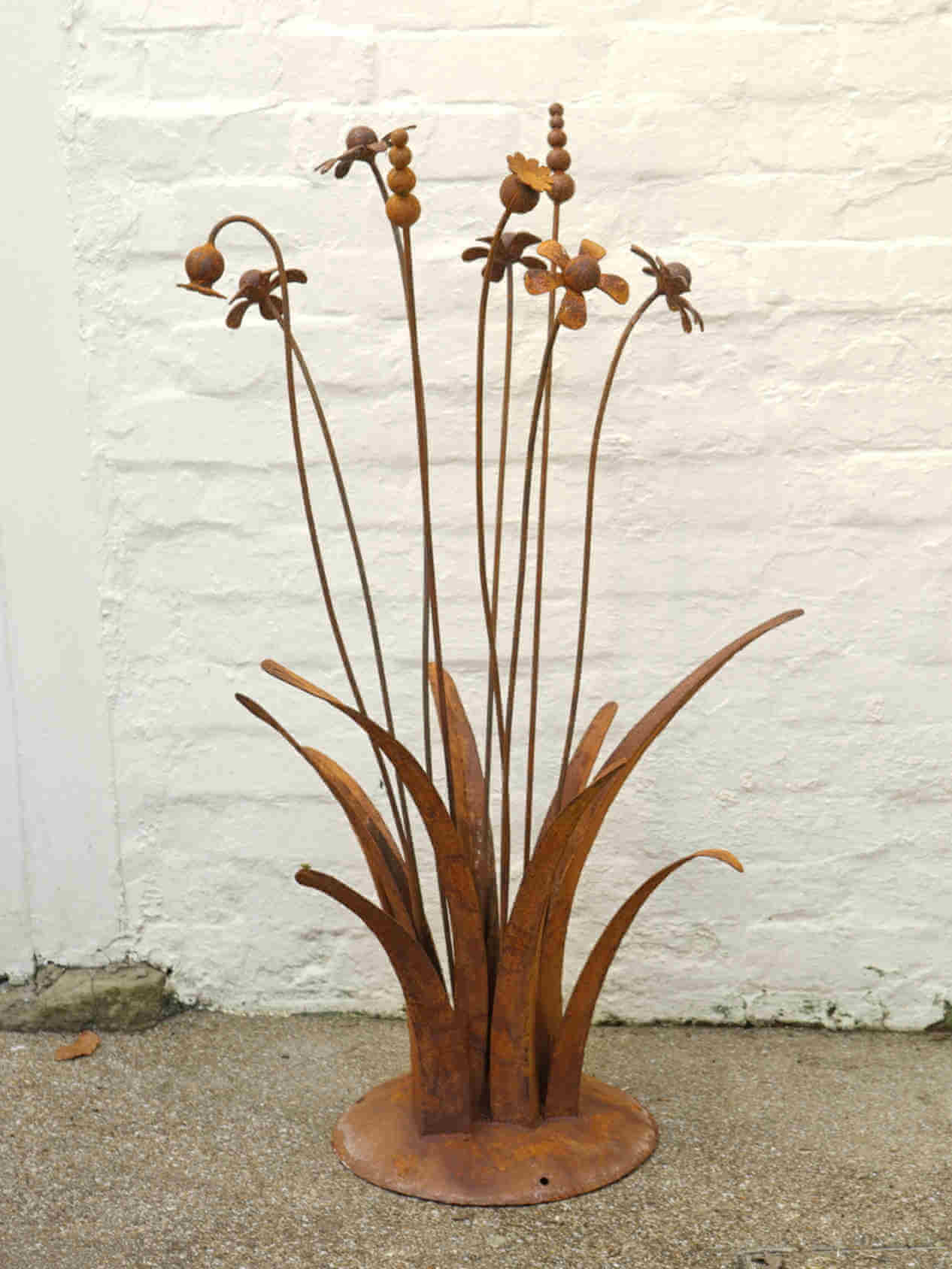 A rusty steel meadow composition - poppies, chamomiles, heleniums and reeds rising through tall grasses on a hand-formed domed base, against a white brick wall.