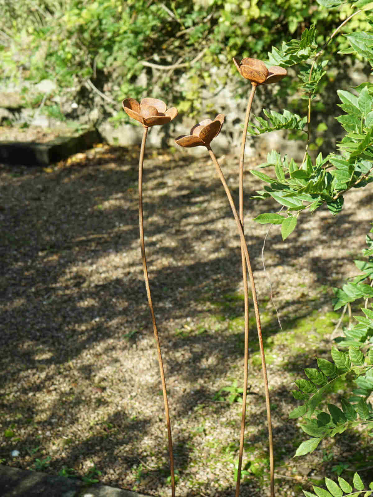 Three rusty metal buttercup sculptures  on slender stems with green leaves in the background