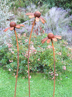 Three rusty metal Chamomile flower sculptures in a garden setting with greenery and flowers.