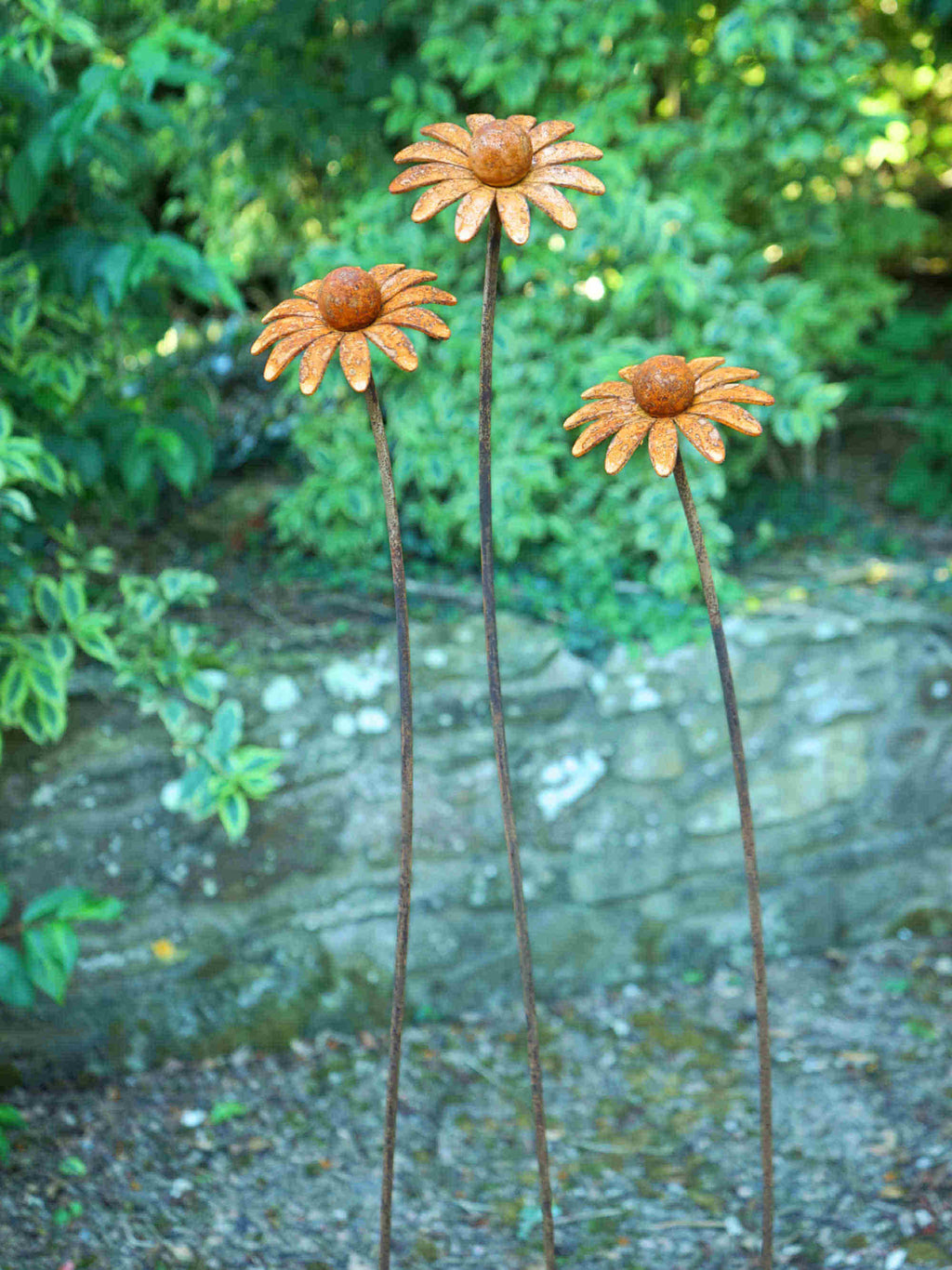 Three rusted metal flower garden stakes shaped like daisies, standing in a garden with green foliage and a stone wall background.