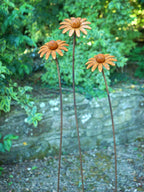 Three rusted metal flower garden stakes shaped like daisies, standing in a garden with green foliage and a stone wall background.