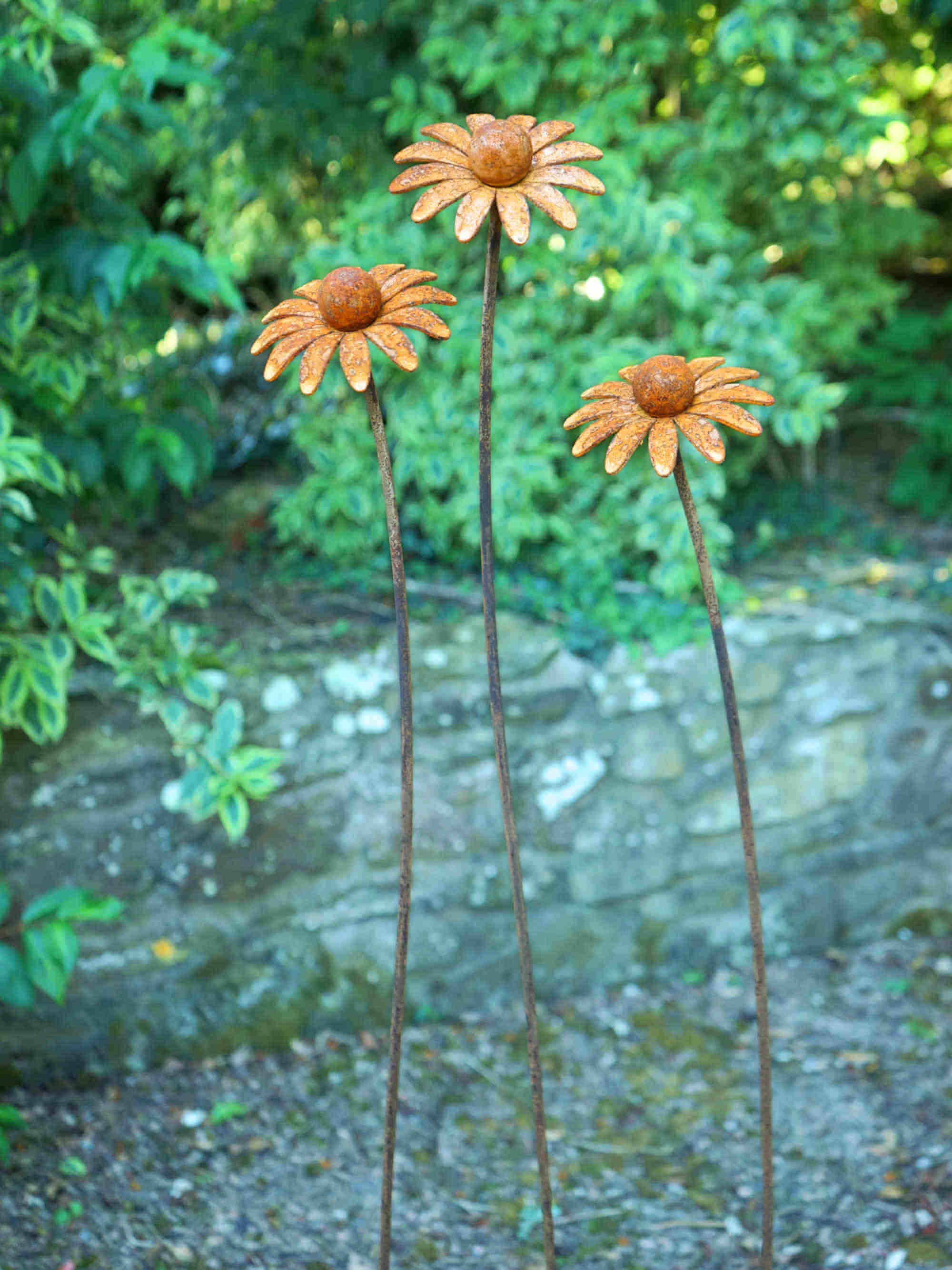 Three rusted metal flower garden stakes shaped like daisies, standing in a garden with green foliage and a stone wall background.