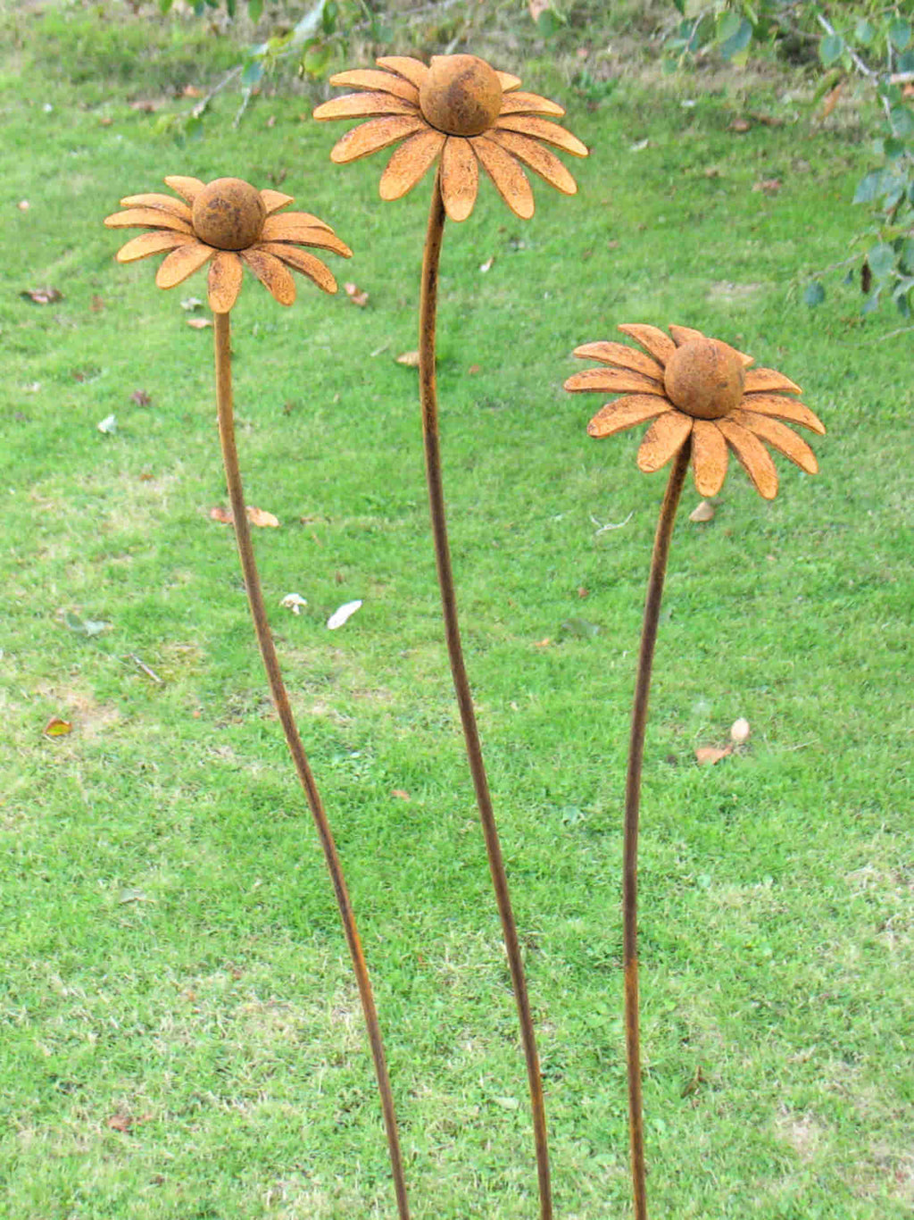 Three rusted metal daisy garden stakes with orange-brown flower heads and long brown stems, set in a green grassy outdoor area.