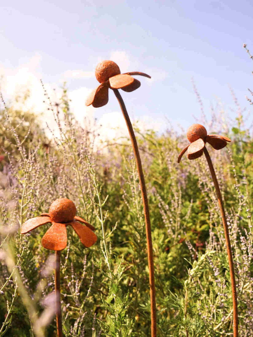 Rusty metal Heleniums flower sculptures in a garden setting with a clear blue sky.