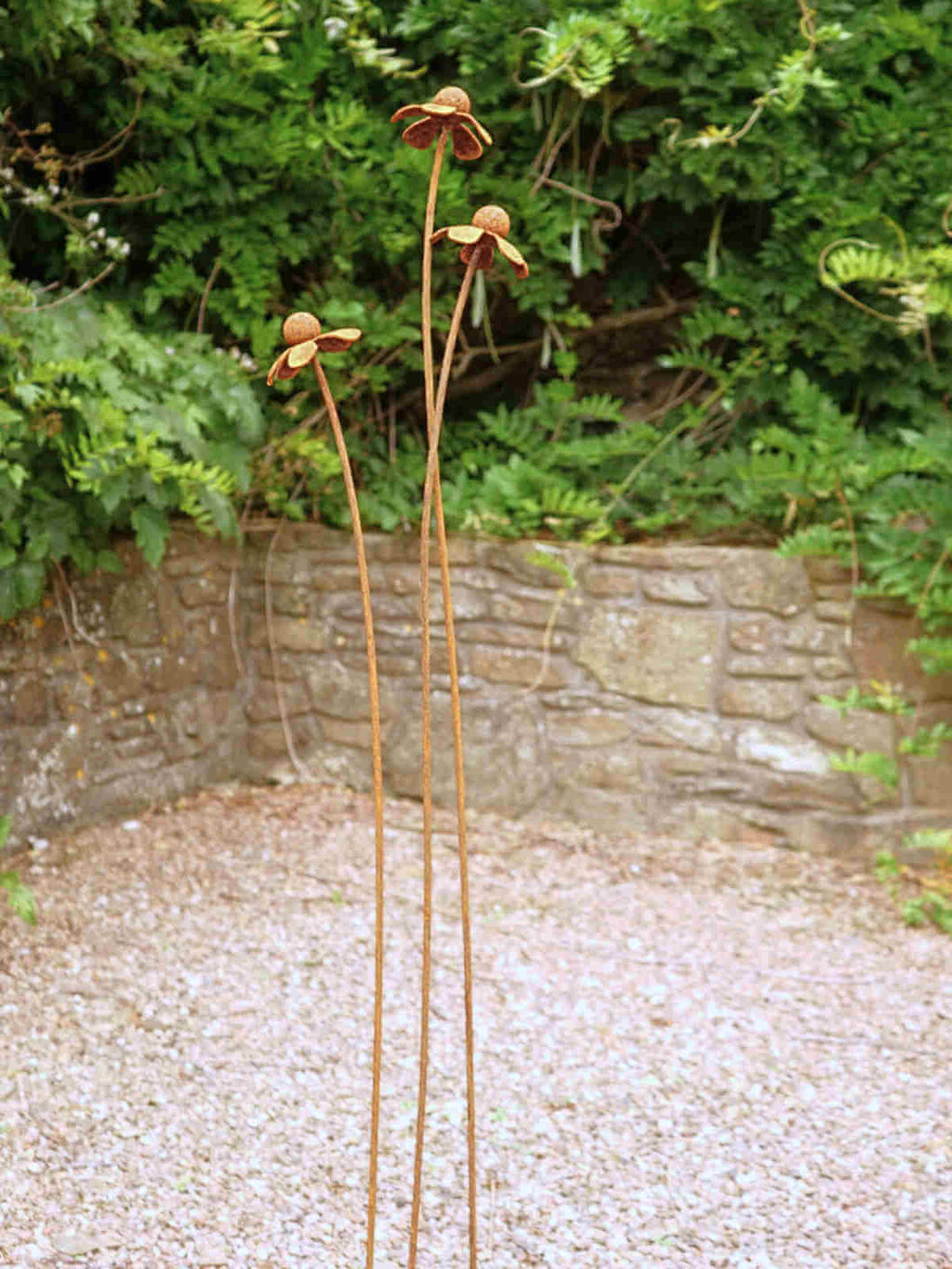 Rusty metal Heleniums flower sculptures on a gravel path with greenery and a stone wall in the background.