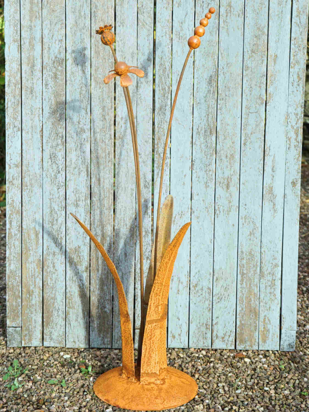 A rusty steel meadow composition - helenium, reed and poppy seed head rising through tall grass stems, freestanding on a domed steel base standing against a weathered wooden background.