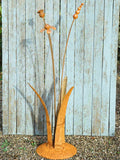 A rusty steel meadow composition - helenium, reed and poppy seed head rising through tall grass stems, freestanding on a domed steel base standing against a weathered wooden background.