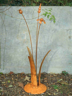 A rusty steel meadow composition - helenium, reed and poppy seed head rising through tall grass stems, freestanding on a domed steel base standing in front of a mottled grey wall.