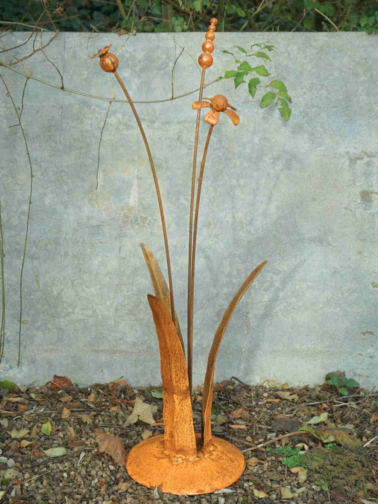 A rusty steel meadow composition - helenium, reed and poppy seed head rising through tall grass stems, freestanding on a domed steel base standing in front of a mottled grey wall.