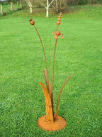 A rusty steel meadow composition - helenium, reed and poppy seed head rising through tall grass stems, freestanding on a domed steel base standing on a grassy lawn.