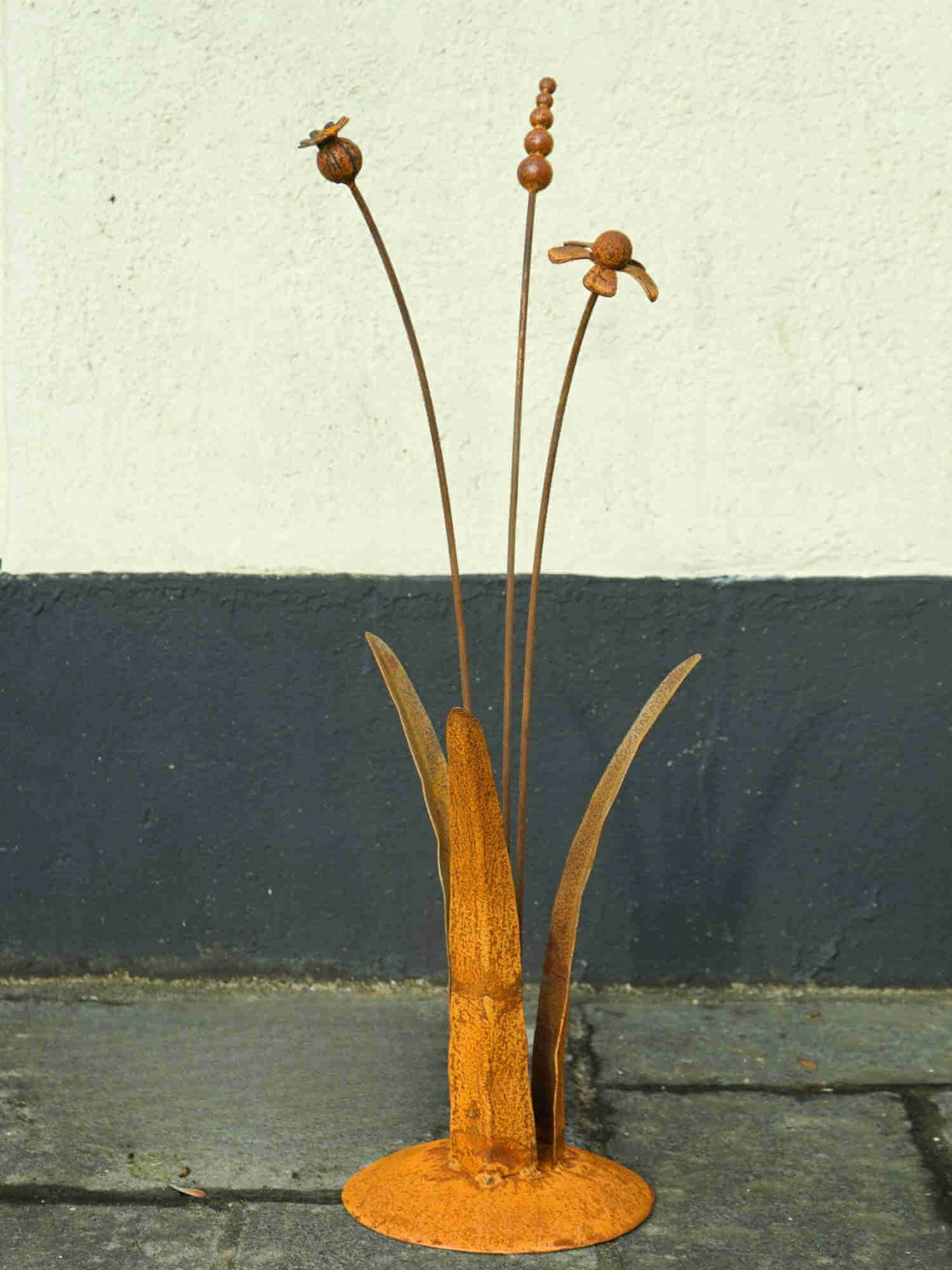 A rusty steel meadow composition - helenium, reed and poppy seed head rising through tall grass stems, freestanding on a domed steel base standing in front of a black and white wall.