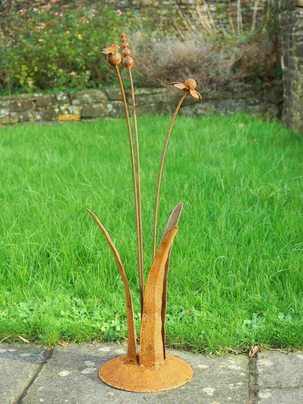 A rusty steel meadow composition - helenium, reed and poppy seed head rising through tall grass stems, freestanding on a domed steel base standing on a garden path.
