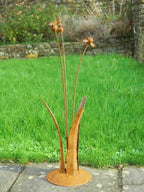 A rusty steel meadow composition - helenium, reed and poppy seed head rising through tall grass stems, freestanding on a domed steel base standing on a garden path.