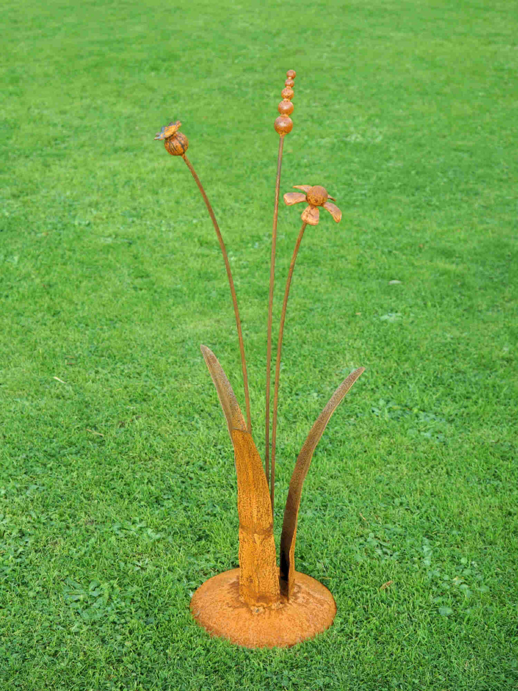 A rusty steel meadow composition - helenium, reed and poppy seed head rising through tall grass stems, freestanding on a domed steel base on a green lawn.