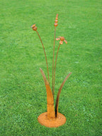 A rusty steel meadow composition - helenium, reed and poppy seed head rising through tall grass stems, freestanding on a domed steel base on a green lawn.