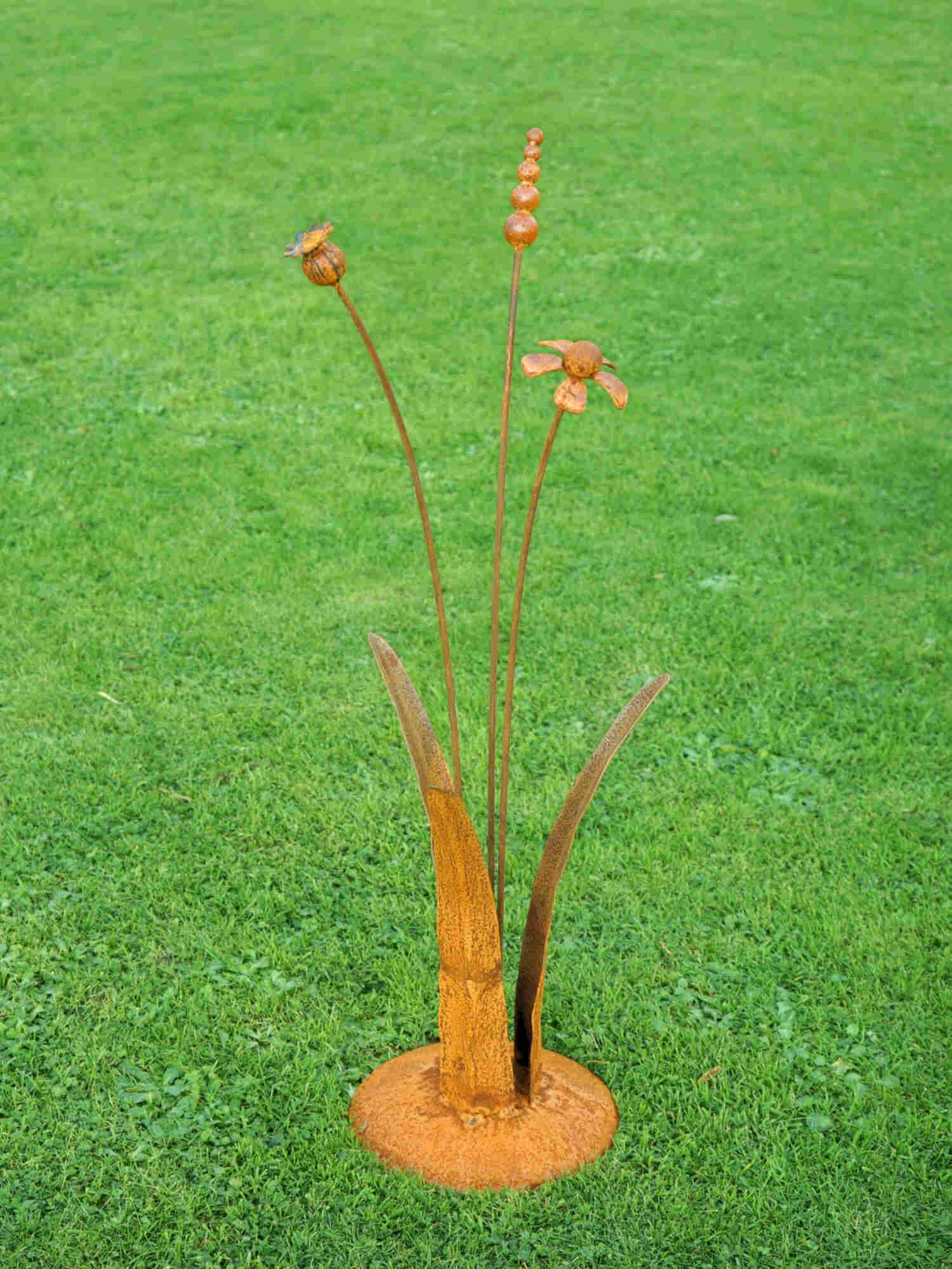 A rusty steel meadow composition - helenium, reed and poppy seed head rising through tall grass stems, freestanding on a domed steel base on a green lawn.