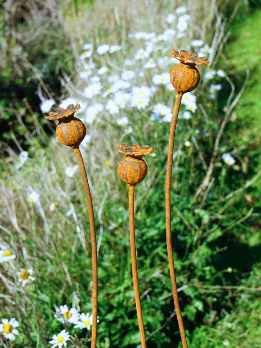 Decorative metal poppy seed head sculptures in a natural setting with greenery and white flowers.