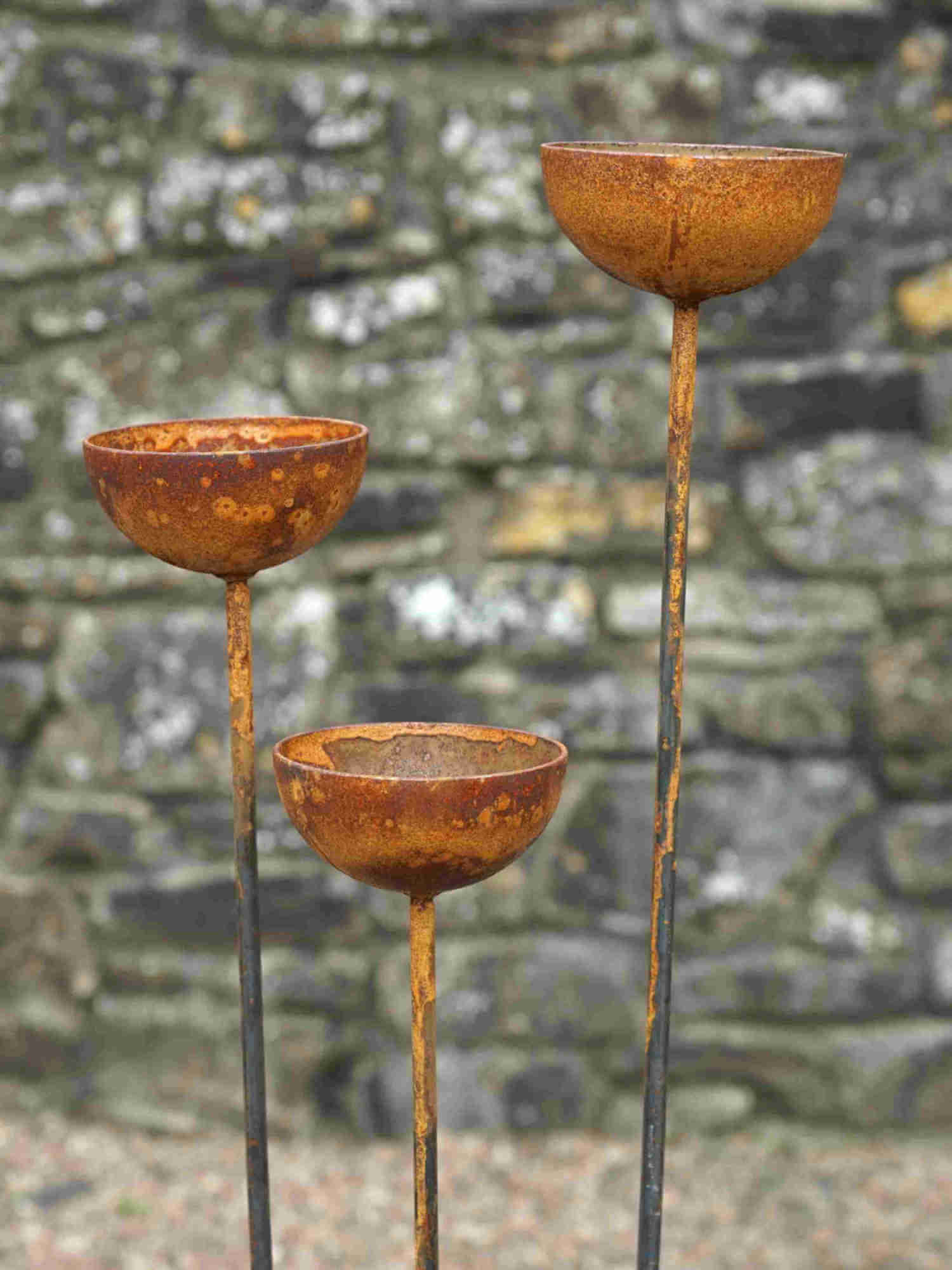 Three rusted metal rain catchers on stands against a stone wall background