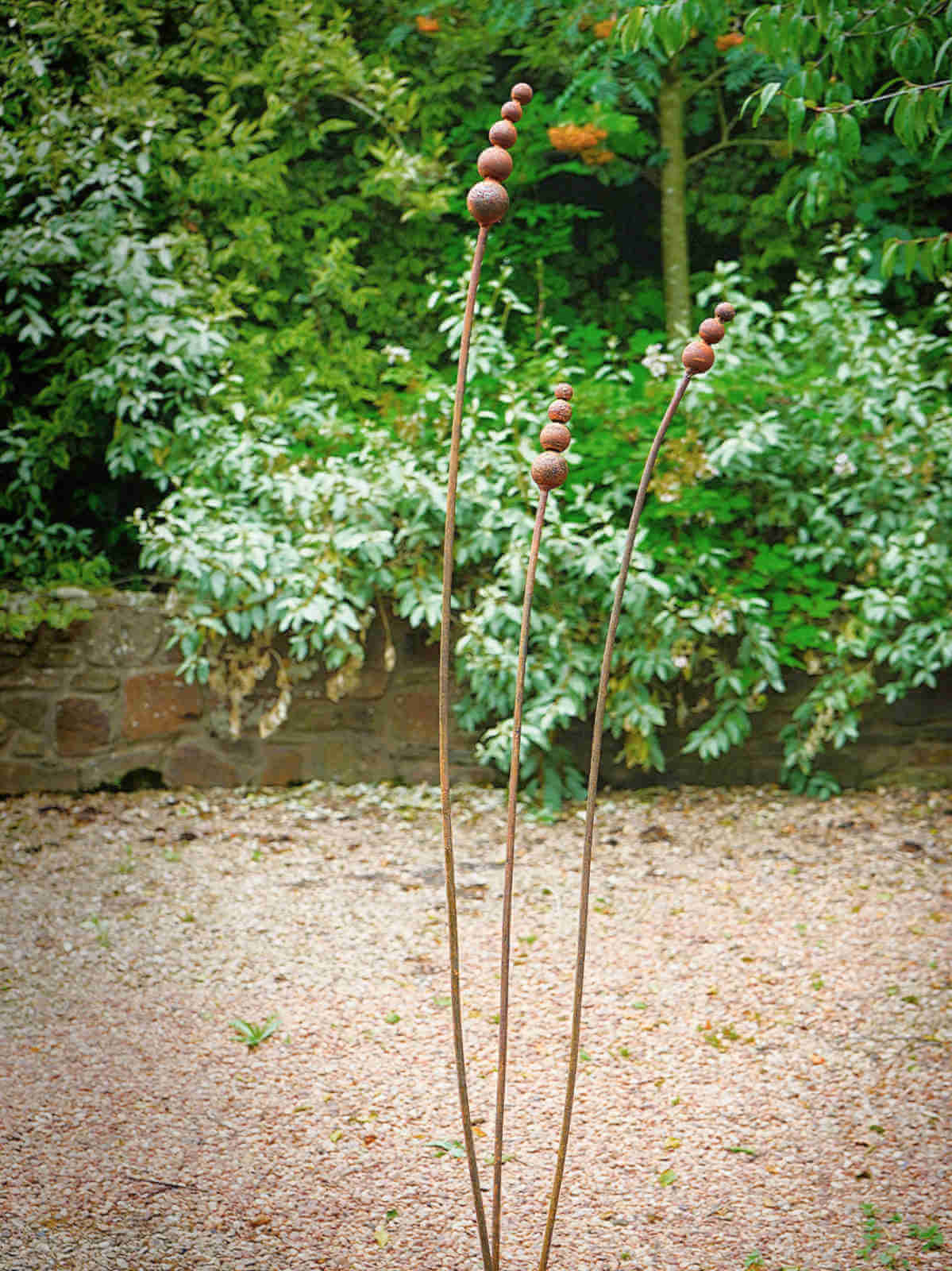 Rusty metal reed garden sculptures on a gravel path with greenery in the background
