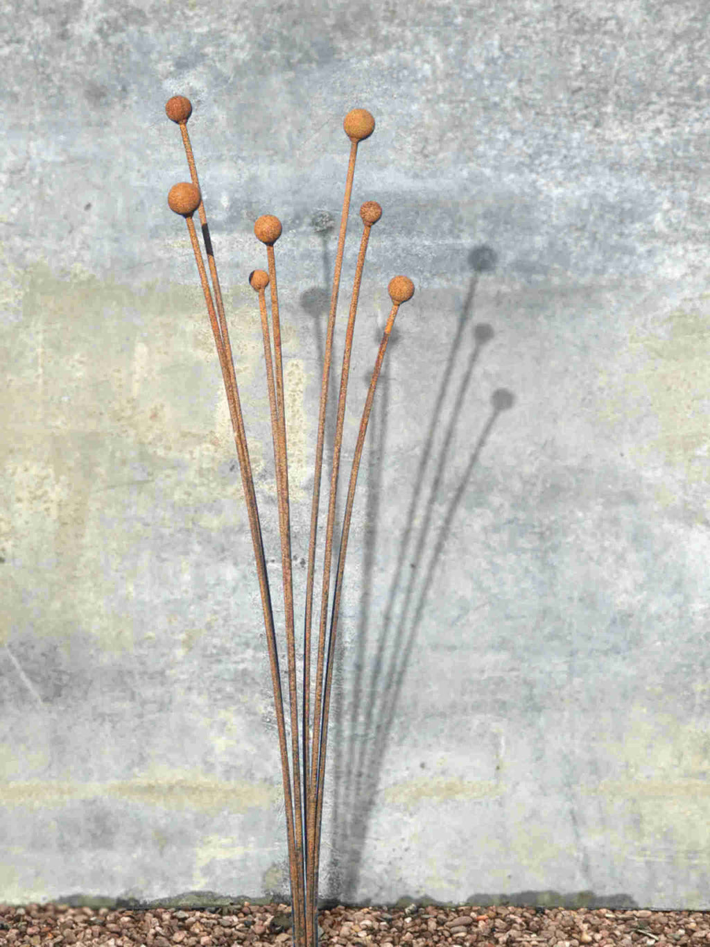 Rusty metal Seed Head garden sculpture  with spherical ends against a textured wall.