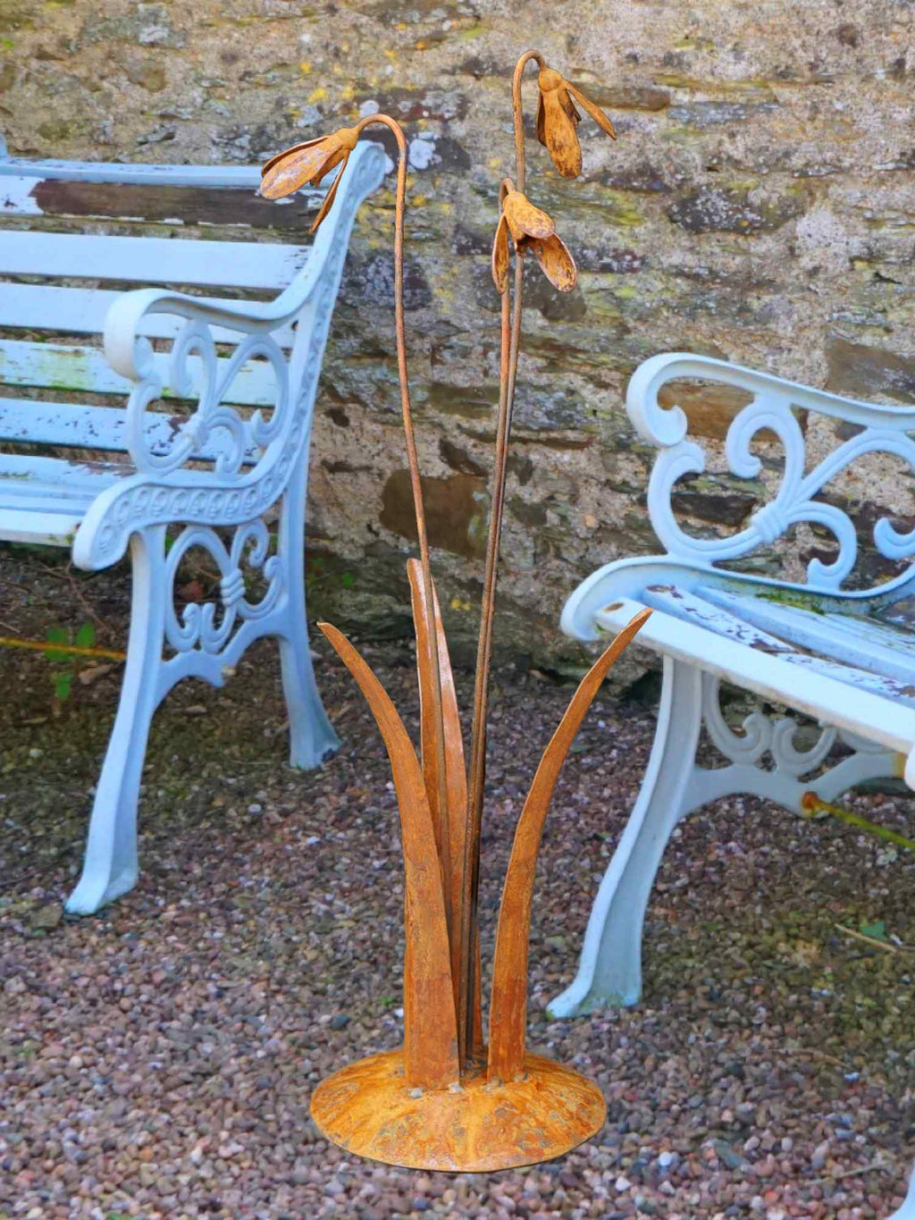 A rusty steel snowdrop composition - three drooping flowers of ascending heights rising through tall grass stems, freestanding on a domed steel base standing on a gravel base with pale blue garden chairs behind.