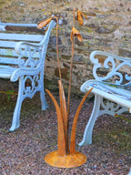 A rusty steel snowdrop composition - three drooping flowers of ascending heights rising through tall grass stems, freestanding on a domed steel base standing on a gravel base with pale blue garden chairs behind.