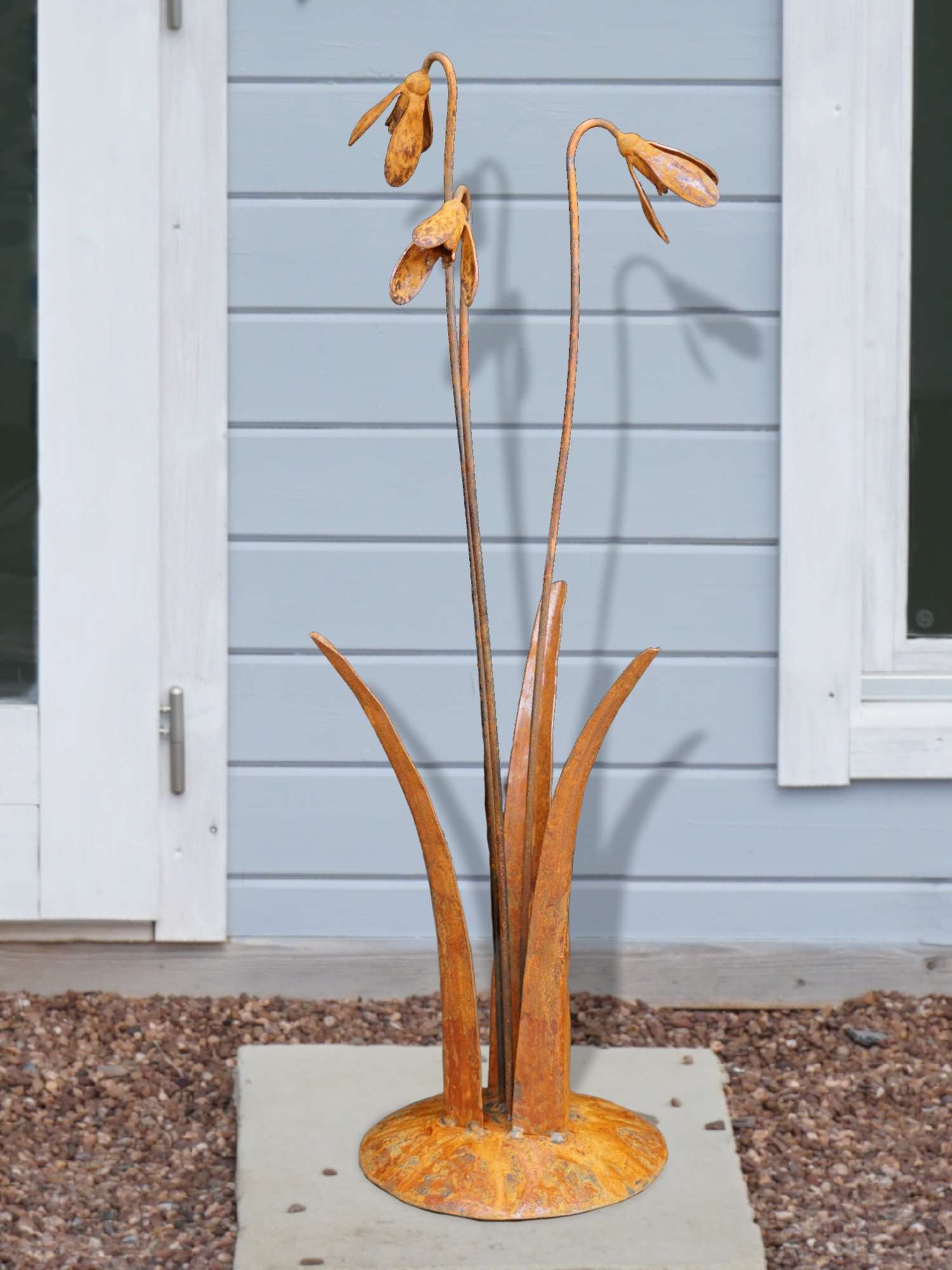 A rusty steel snowdrop composition - three drooping flowers of ascending heights rising through tall grass stems, freestanding on a domed steel base standing in front of a wooden cabin.