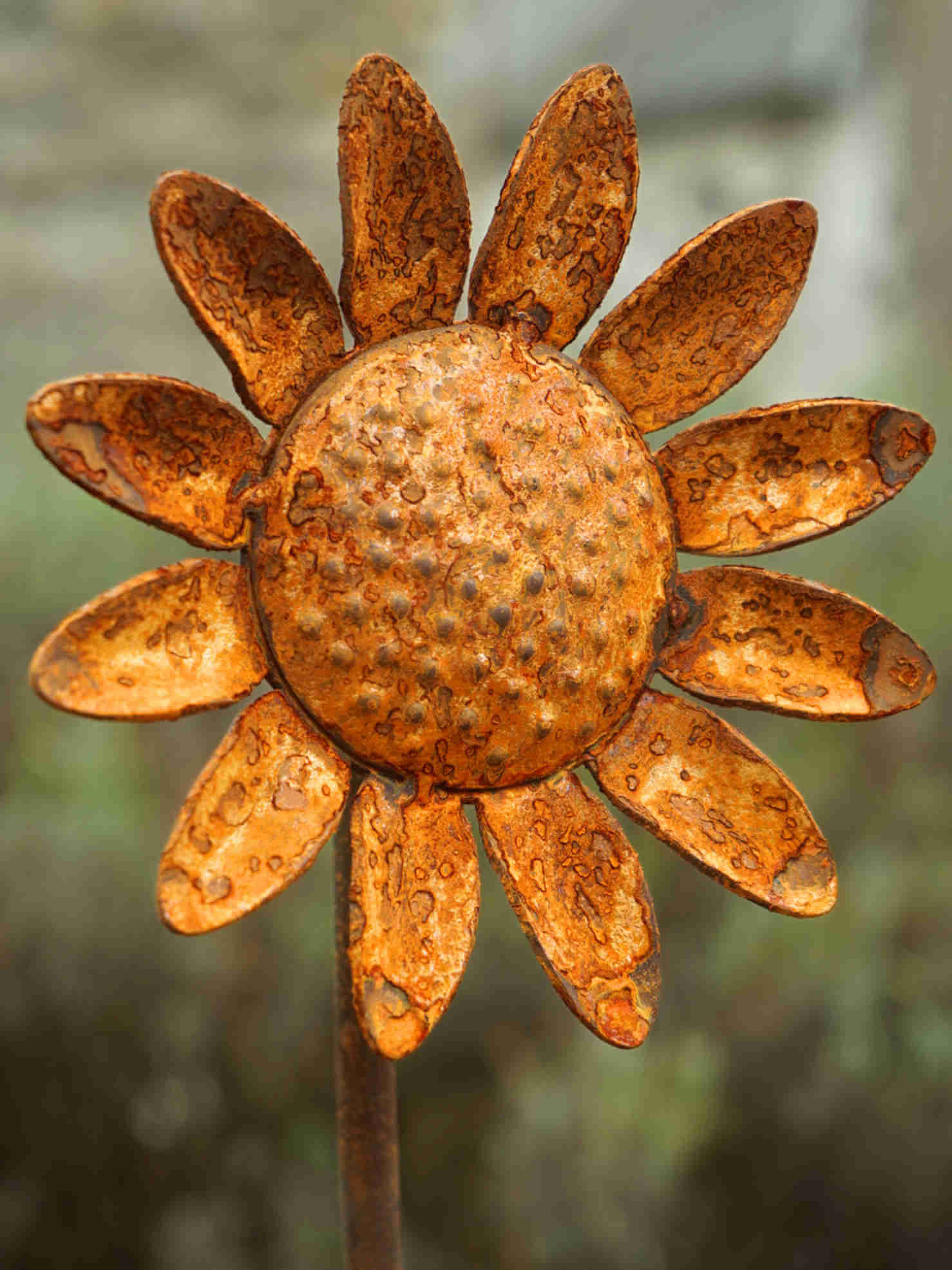 A rusted metal sunflower garden stake decoration with textured, weathered orange-brown petals and a dotted center, set against a blurred green garden background.