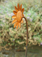 A rusted metal garden sculpture shaped like a sunflower on a long stem, photographed outdoors against a blurred green and beige garden background.