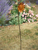 A rusted metal garden stake shaped like a sunflower stands in a grassy lawn with blurred flowers and lavender in the background.