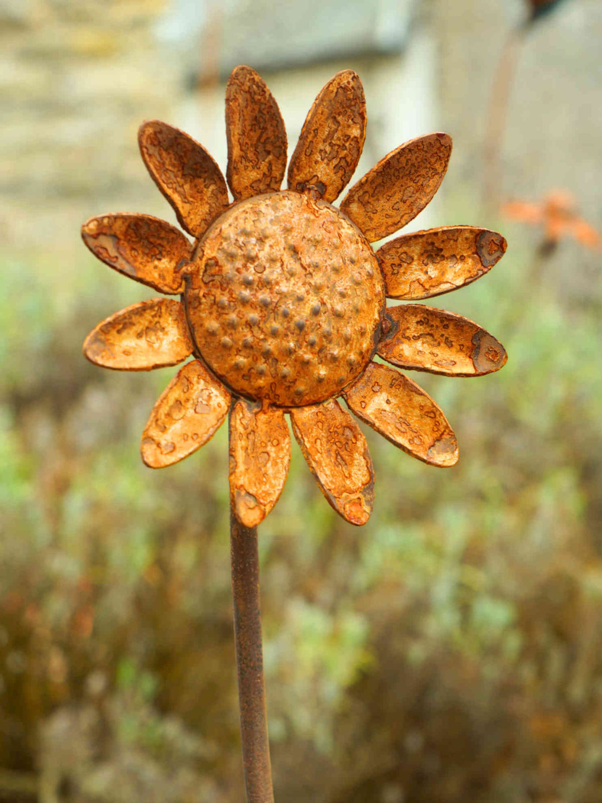A rustic metal sunflower garden stake with a textured, rusted orange-brown finish, photographed outdoors against a blurred natural background.