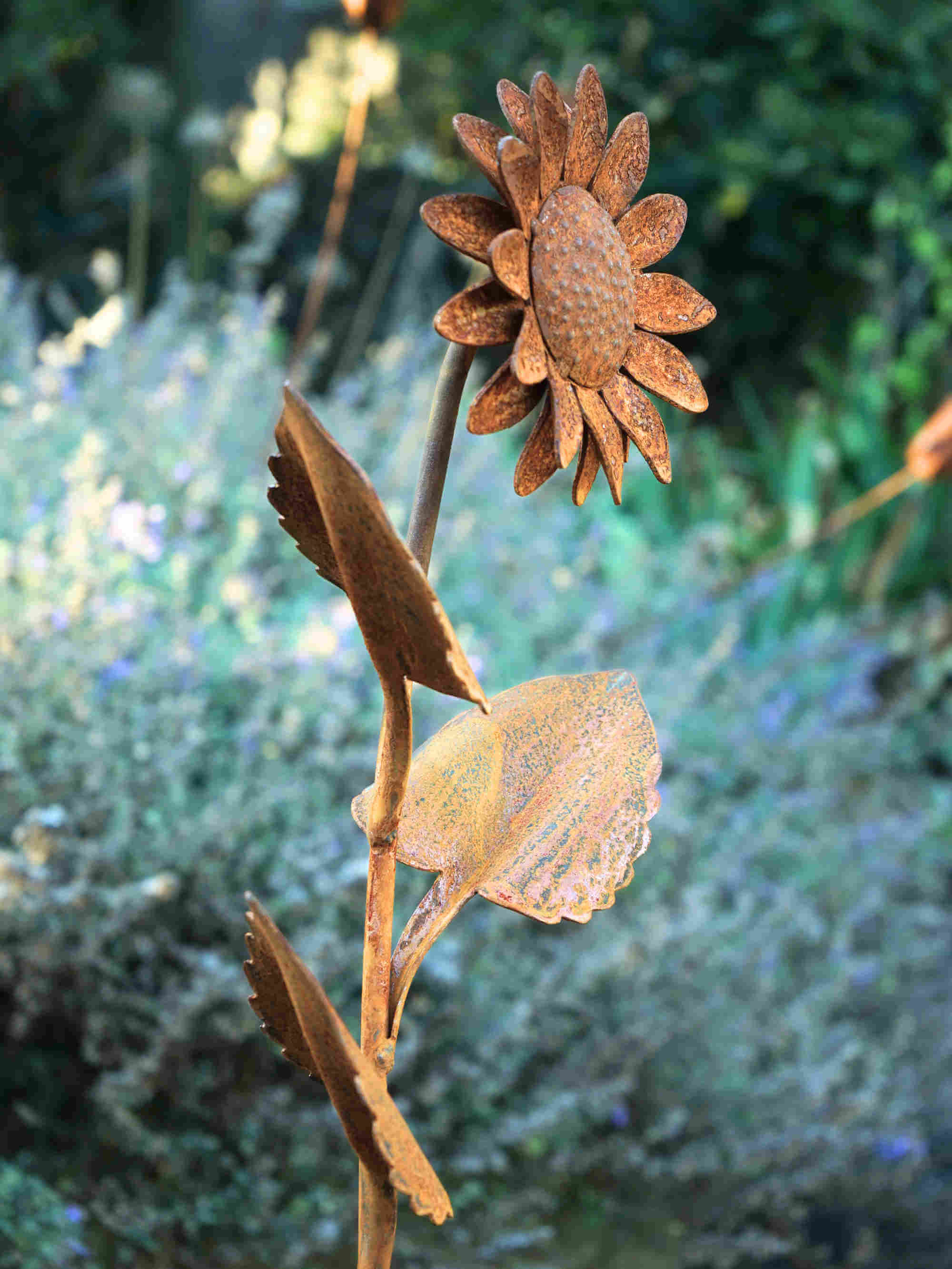 Rustic metal garden sculpture of a sunflower with three leaves, shown outdoors against a blurred green garden background.