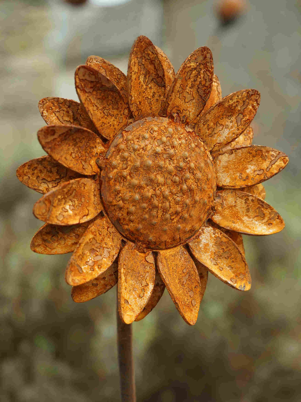 Rustic metal sunflower garden stake with textured, weathered orange-brown petals and a detailed centre, shown in a blurred outdoor setting.