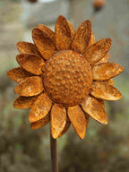 Rustic metal sunflower garden stake with textured, weathered orange-brown petals and a detailed centre, shown in a blurred outdoor setting.
