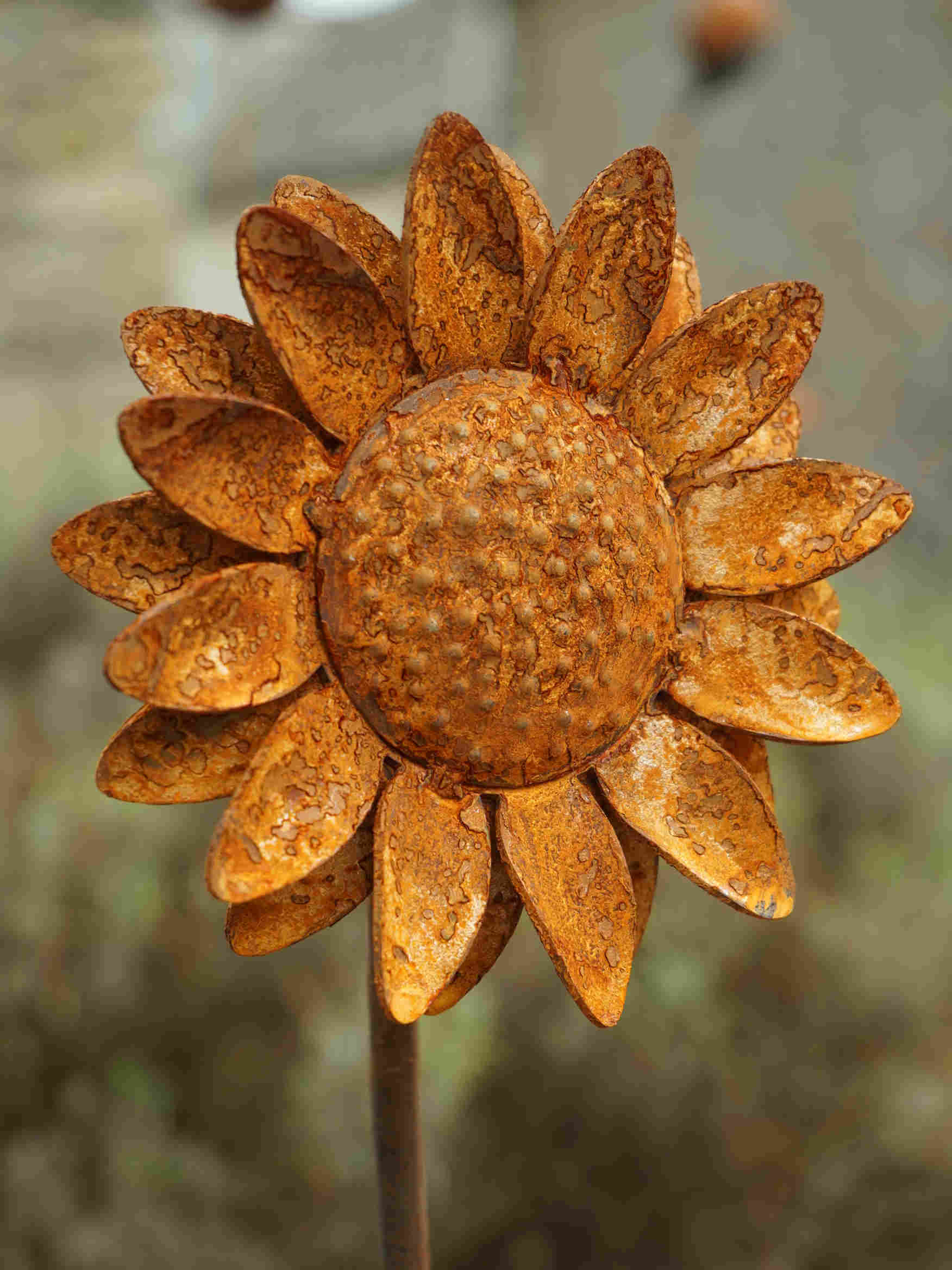 Rustic metal sunflower garden stake with textured, weathered orange-brown petals and a detailed centre, shown in a blurred outdoor setting.