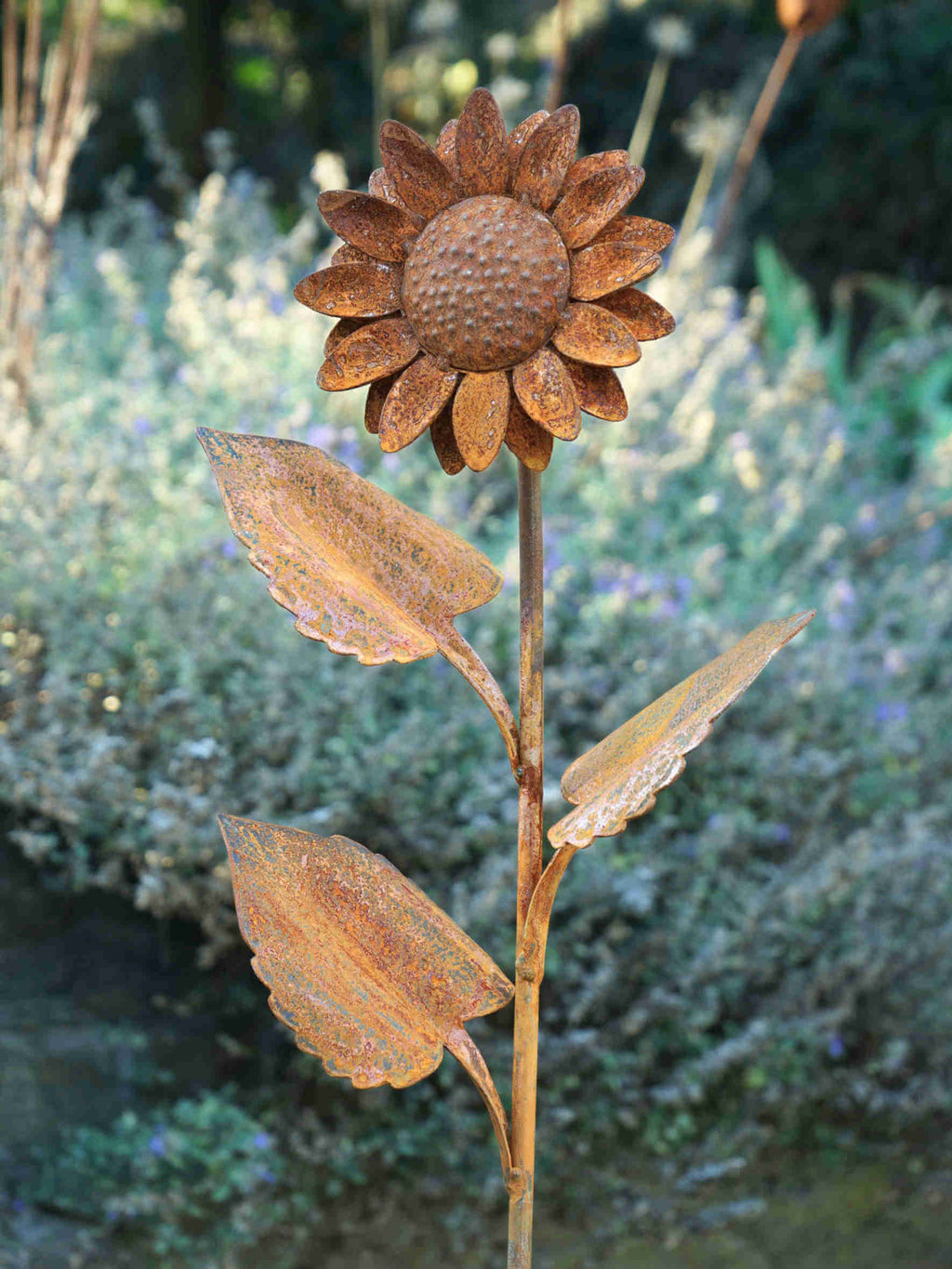 Rustic metal garden sculpture of a sunflower with textured brown petals and leaves on a garden stake, photographed outdoors against a blurred green and purple garden background.