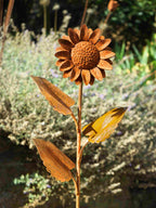 Rustic metal garden sculpture of a sunflower with textured petals, a detailed centre, and three leaves on a curved stem, photographed outdoors against a blurred garden background.