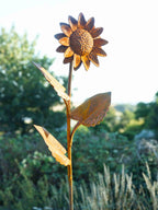 Rustic metal garden sculpture of a single sunflower with textured petals, a detailed centre, and three leaves on a curved stem, photographed outdoors against a blurred green garden background.