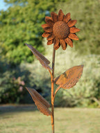Rustic metal garden sculpture of a single sunflower with textured petals, a detailed center, and three leaves on a stem, photographed outdoors against a blurred green garden background.